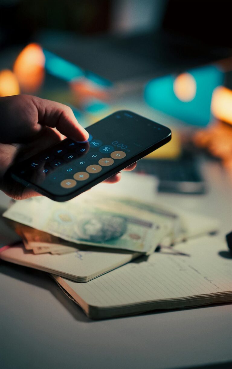 A hand holds a smartphone running a calculator app, with cash and a notebook on a desk, symbolizing budgeting and finance management.
